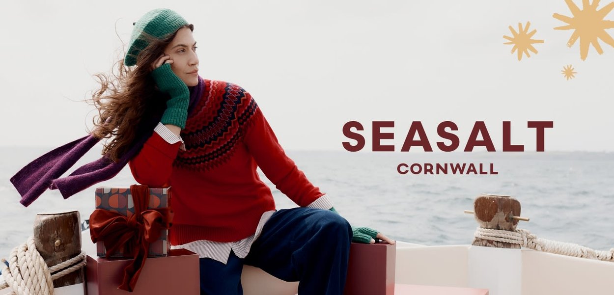 Roter Pullover mit gemustertem Schulterbereich, grüne Handschuhe und kariertes Hemd. Lila Schal, lockere marineblaue Hose. Geschenke mit Schleife auf einem Boot. Ozean im Hintergrund.