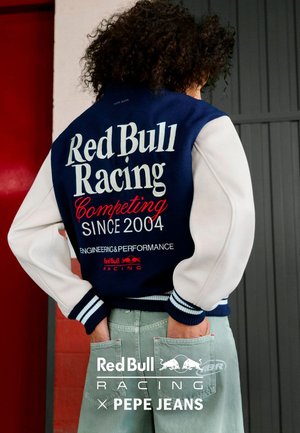 Persona con cabello rizado que lleva una chaqueta azul marino y blanca con el texto "Red Bull Racing Competing Since 2004", con las manos en los bolsillos traseros de unos pantalones verde claro.
