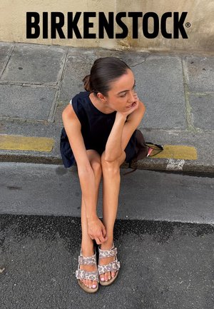 Frau sitzt auf einem Trottoirrand und trägt Birkenstock-Sandalen mit Leopardenmuster sowie ein schwarzes ärmelloses Kleid auf einem städtischen Gehweg.