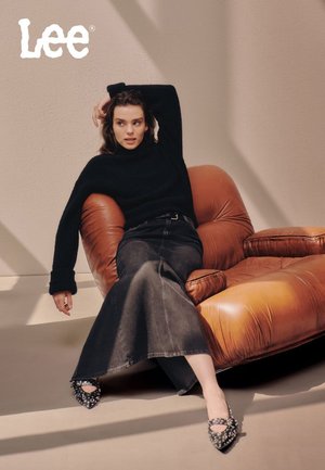 Svart stickad tröja, mörk denim maxi kjol och spikade svarta klackar. Sitter på en brun lädersoffa med en texturerad yta.