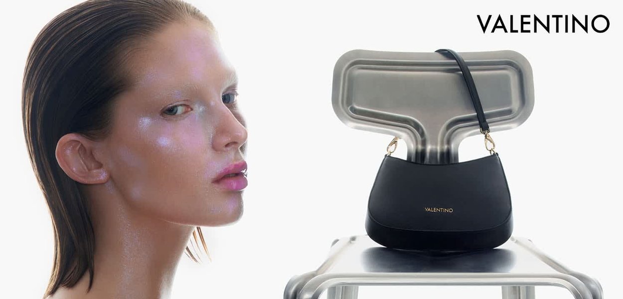 Modella con pelle luminosa e capelli lisci tirati indietro accanto a una borsa nera Valentino su una sedia metallica contro uno sfondo bianco.