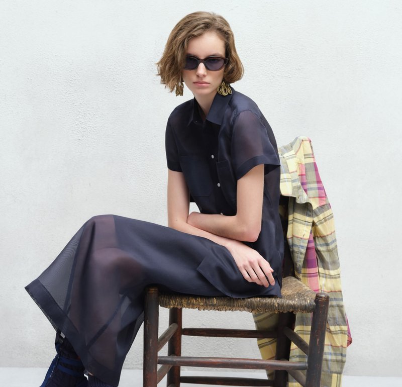 Vrouw met kort haar, gekleed in een donkere doorschijnende jurk en zonnebril, zittend op een gevlochten stoel met een geruit jasje over de rugleuning.