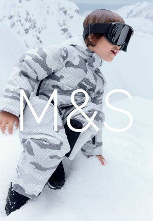 Dziecko w szarym kombinezonie zimowym w kamuflażu i czarnych goglach narciarskich kuca na śniegu na tle zaśnieżonych gór.