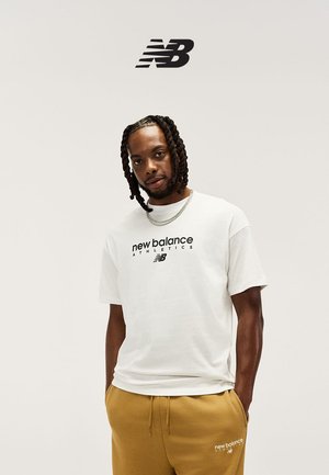 Mann mit geflochtenem Haar, der ein weißes New Balance Athletics T-Shirt und senfgelbe Jogginghosen trägt, Hände in den Taschen, neutraler Hintergrund.