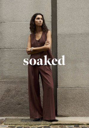 Frau mit lockigem Haar, die einen ärmellosen braunen Overall und schwarze Schuhe trägt, steht mit verschränkten Armen an einer Steinmauer. Der Text lautet "soaked."