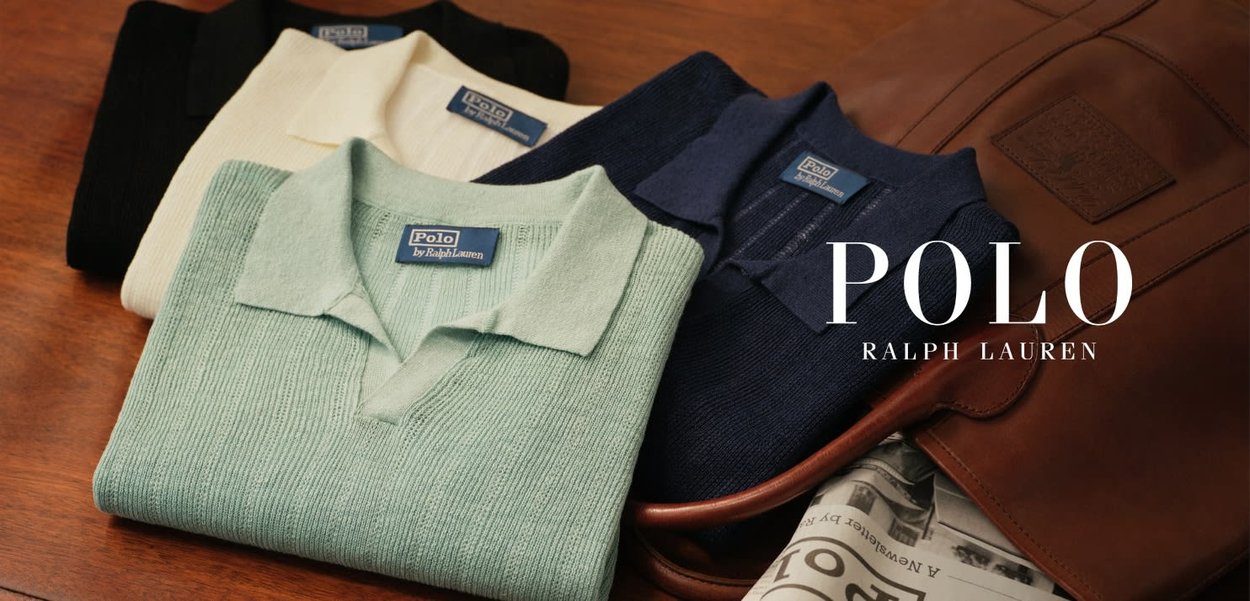 Tre brettede Polo Ralph Lauren gensere i krem, lysegrønn og marineblå på en treflate ved siden av en brun skinnveske og en avis.