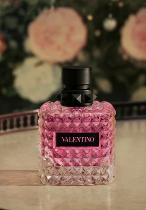 Glass perfume bottle filled with pink liquid labeled Valentino, placed on a marble surface with blurred floral background.