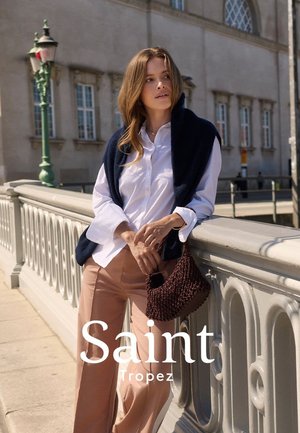 Una modella indossa una sciarpa blu navy sopra una camicia bianca e pantaloni palazzo beige chiaro, tenendo una borsa intrecciata marrone testurizzata, sullo sfondo di una città.