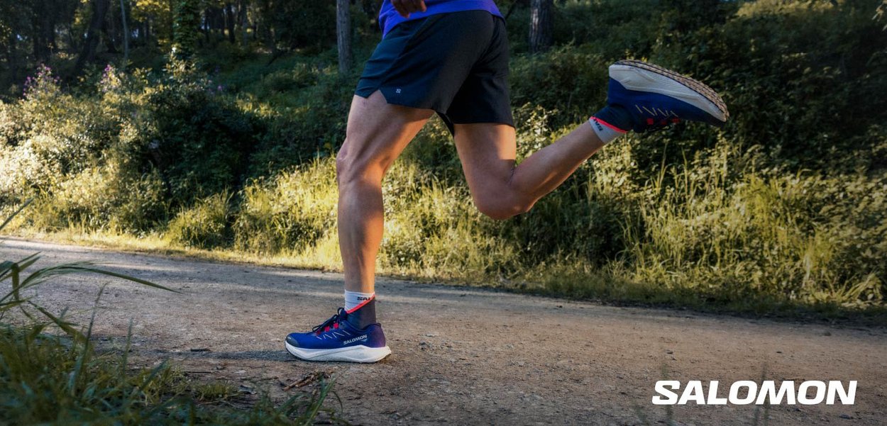 Des chaussures de sport bleues avec des accents rouges, au design léger et en matériau respirant, portées par un coureur sur un chemin en terre entouré de verdure.