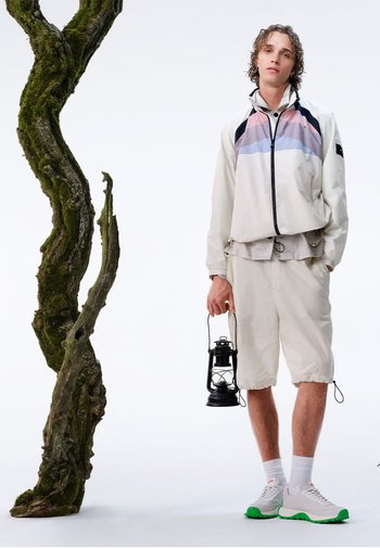 Lightweight cream jacket with multicolor accents, paired with beige shorts. White sneakers with green soles. Holds a black lantern.