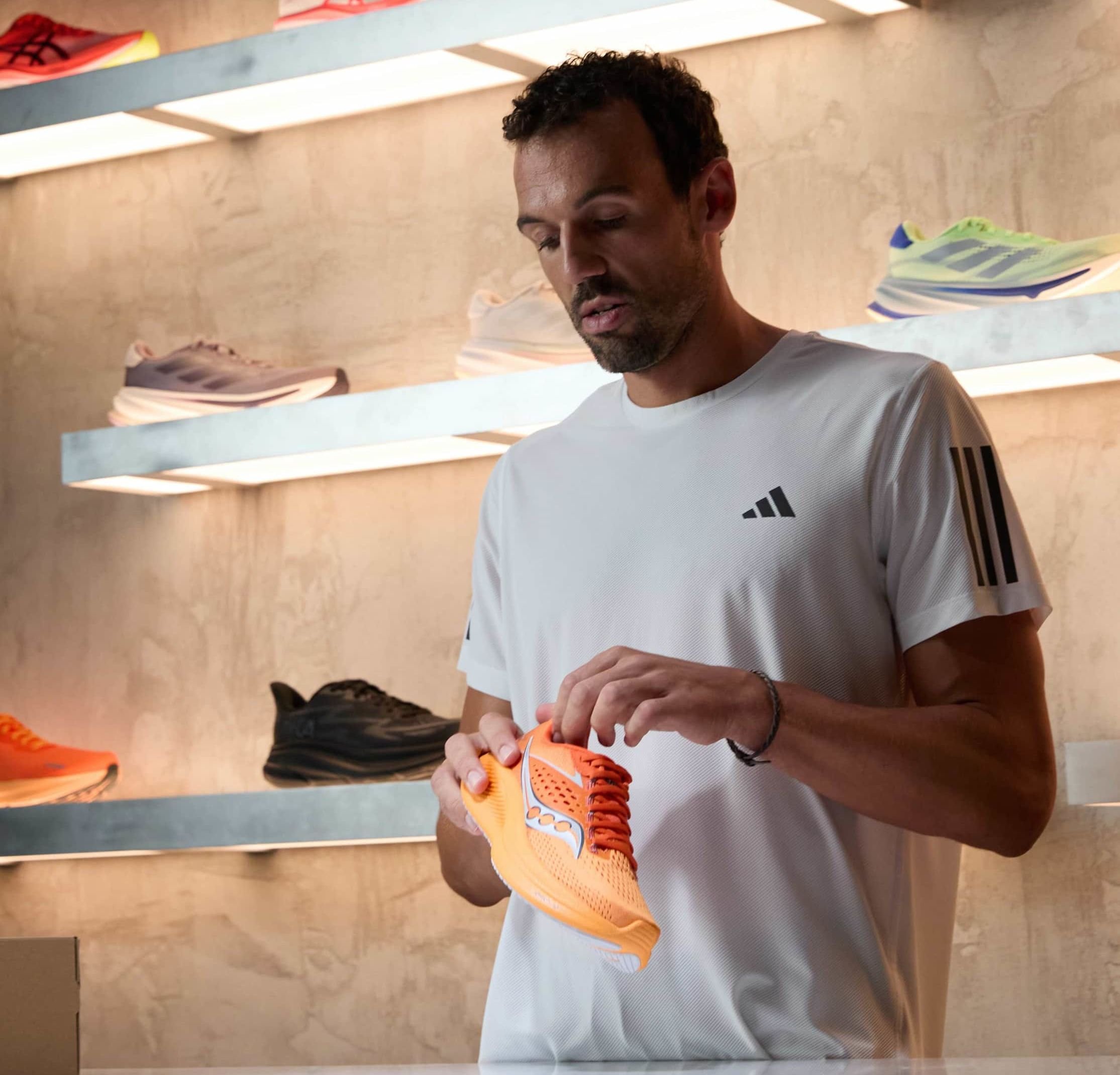 Oranger Laufschuh mit einem Obermaterial aus Mesh, weißen Akzenten und orangenen Schnürsenkeln. Auf einem Regal mit anderen Sportschuhen im Hintergrund präsentiert.