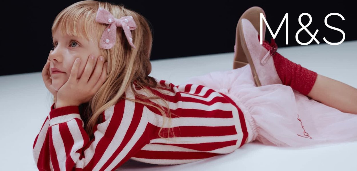 Jeune fille allongée sur le ventre, portant un pull rayé rouge et blanc, une jupe rose, des chaussures roses et un nœud rose dans les cheveux.