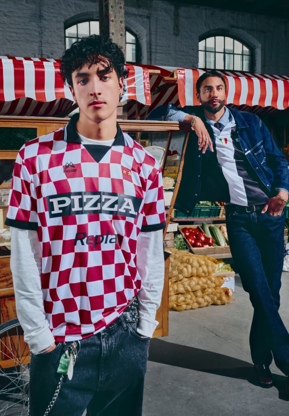Maglia a scacchi rossi e bianchi con logo "PIZZA", colletto nero; modello in jeans neri strappati, catena d'argento. Ambientazione di mercato con prodotti freschi.