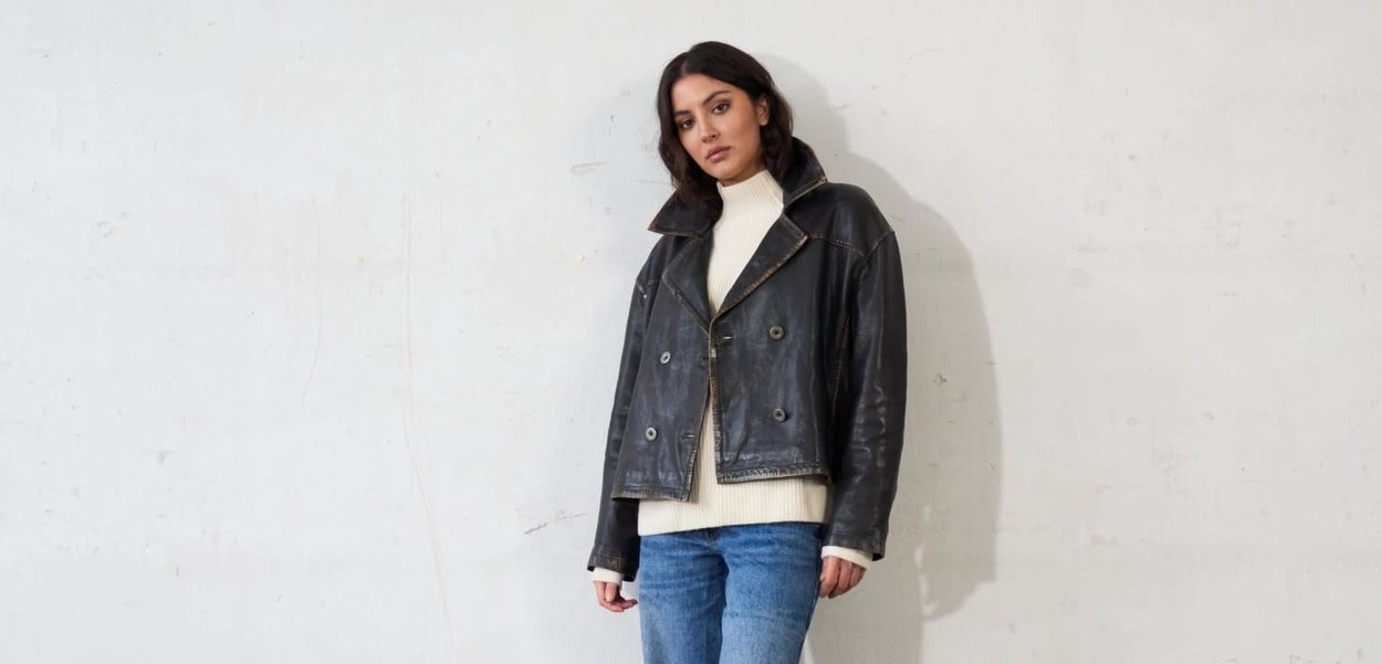 Giacca corta in pelle nera con ampio colletto, bottoni doppiopetto e cuciture a contrasto. Indossata sopra un maglione bianco a maglia.