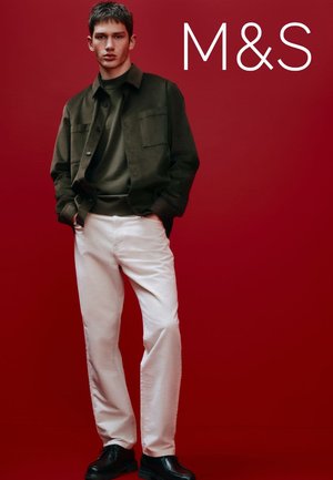 Veste vert foncé sur un pull assorti, pantalon blanc et chaussures noires. Le mannequin se tient devant un fond rouge. Look décontracté et moderne.