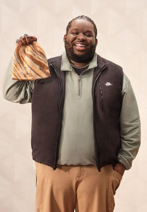 Braune Fleeceweste über einem grünen Langarmshirt. Die Person hält eine marmorierte orange-goldene Mütze. Silberne Kettenverzierung und kurze Haare.