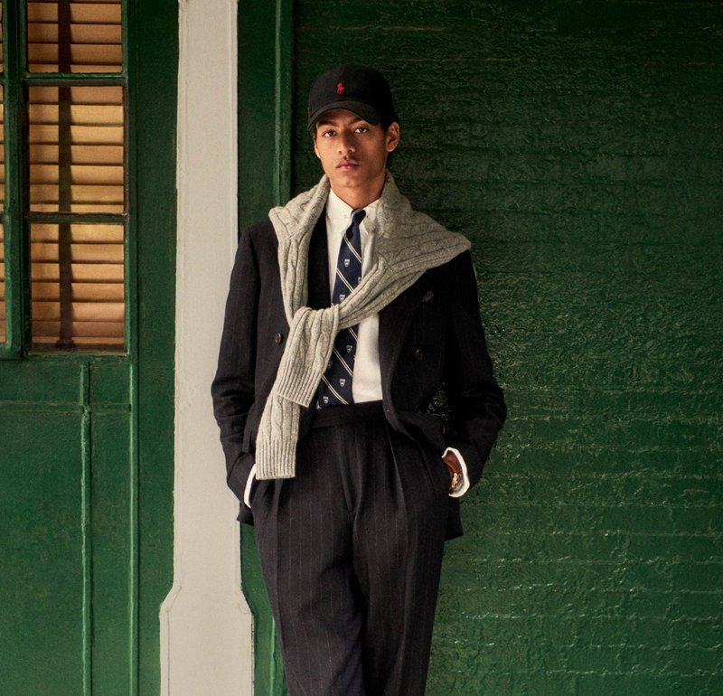 Completo a doppio petto in tessuto a righe, camicia bianca, cravatta blu navy, maglione grigio a treccia appoggiato sulle spalle, berretto nero. Sfondo verde.