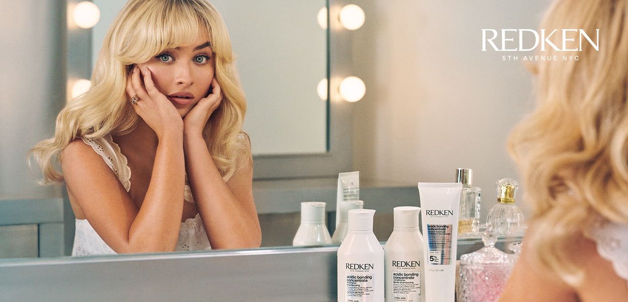 Redken hair care products in white bottles with black labels on a mirrored vanity. Blond hair, soft waves, and neutral background lighting.