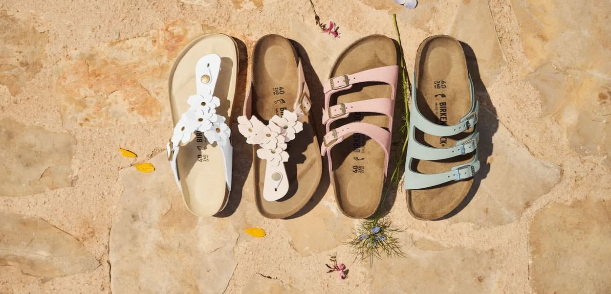 Cuatro pares de sandalias Birkenstock en blanco, beige, rosa y azul claro dispuestas en fila sobre una superficie de piedra con pequeñas flores dispersas.