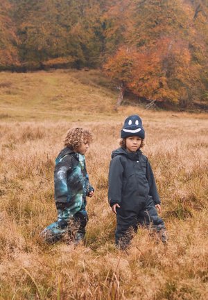 Two children in outerwear: one in a galaxy-patterned jumpsuit, the other in solid dark jumpsuit. Both wear hats. Grass and trees in background.