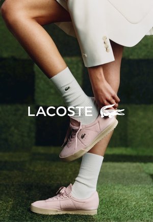 Rosa Leder-Sneaker mit weißen Gummisohlen, kombiniert mit weißen Socken, die ein grünes Logo tragen. Hintergrund mit einer grasähnlichen Textur in Grün.