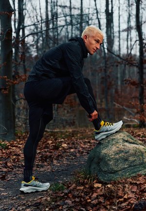 Persoon met donker sportkledij en blond haar die een hardloopschoen strikt op een grote rots op een met bladeren bedekt bospad.