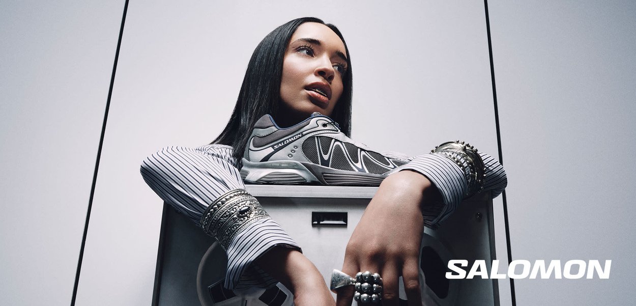 Femme appuyant les bras sur une machine à laver avec une chaussure Salomon posée dessus, portant des bagues et des bracelets, fond blanc minimaliste.