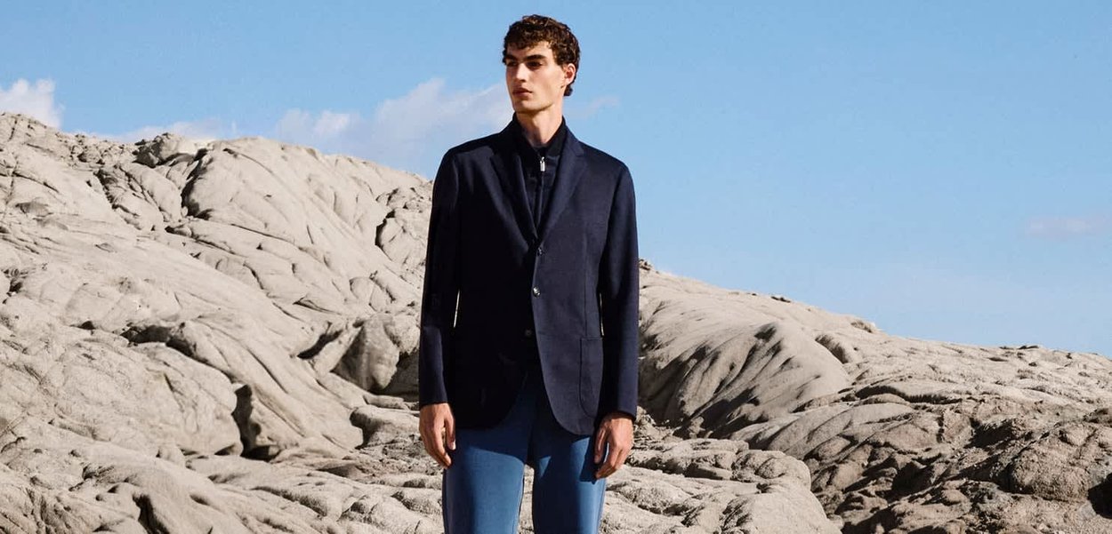 Giovane uomo in blazer blu navy e pantaloni blu che sta su un terreno roccioso sotto un cielo azzurro limpido.