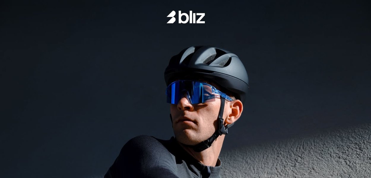 Uomo con casco da ciclismo nero e occhiali da sole sportivi blu su sfondo scuro con logo Bliz sopra.