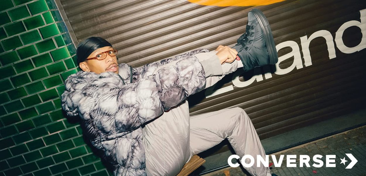 Sneakers alte nere; giacca imbottita grigia con motivo tie-dye; pantaloni grigio chiaro; sfondo piastrellato verde; segnaletica nera e gialla.