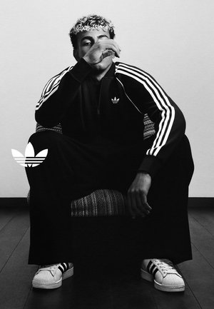 Młoda osoba o kręconych włosach siedzi na krześle, ubrana w czarny dres Adidas i białe trampki z trzema paskami.