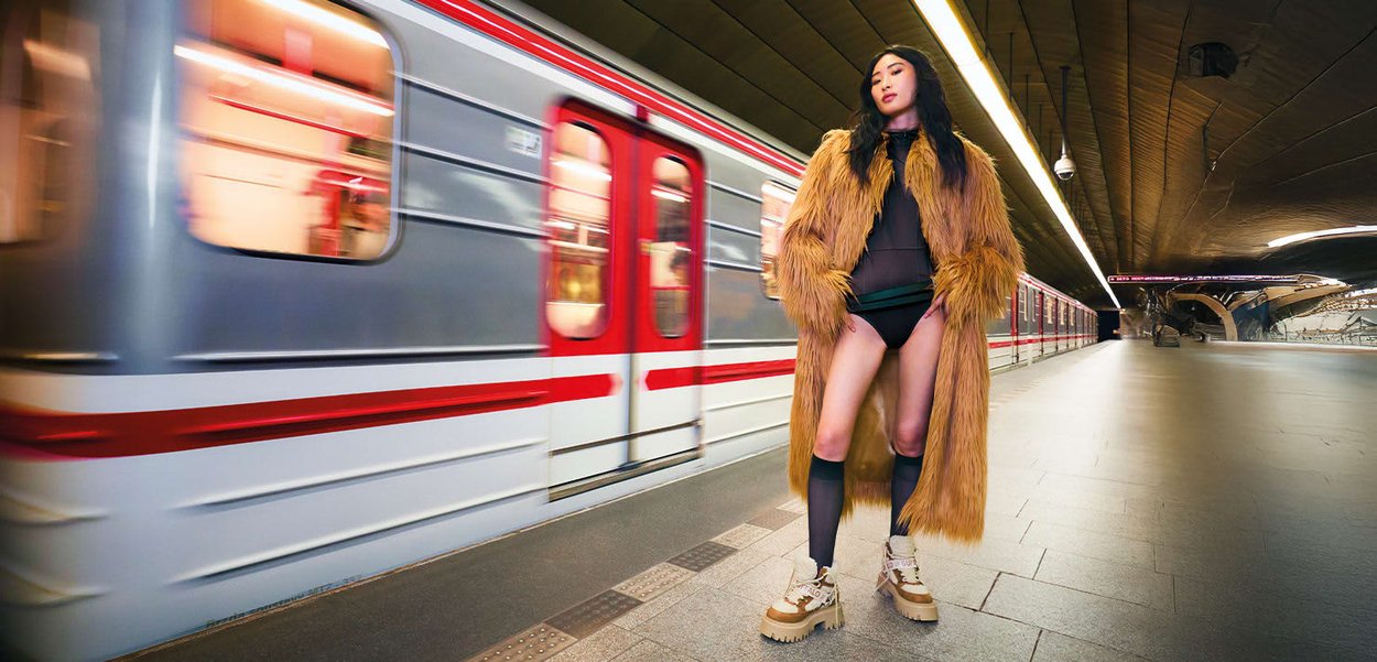 Una giovane donna in un lungo cappotto di pelliccia e stivali si trova su una banchina della metropolitana con un treno in movimento sullo sfondo.