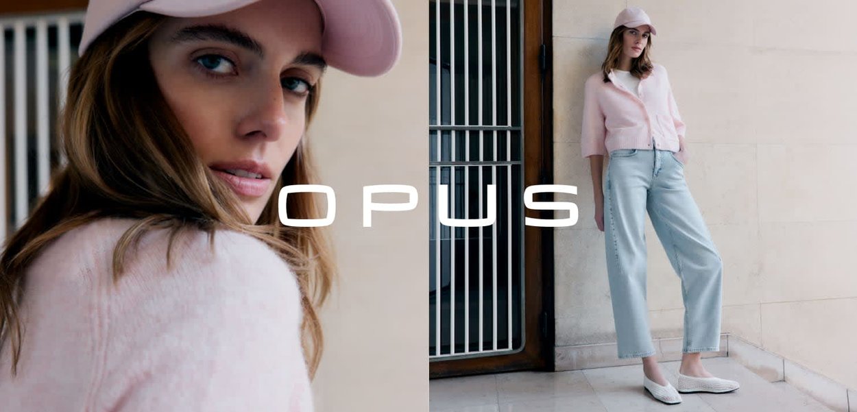 Vrouw in pastelroze vest en pet, gecombineerd met lichtblauwe jeans en witte schoenen, staand tegen een beige stenen muur nabij een metalen poort.