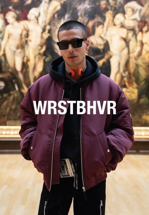 Bordowa kurtka bomberka na czarnym hoodie, czarne spodnie i pomarańczowe słuchawki. Model nosi ciemne okulary przeciwsłoneczne, a na widoku widać torbę na pas.