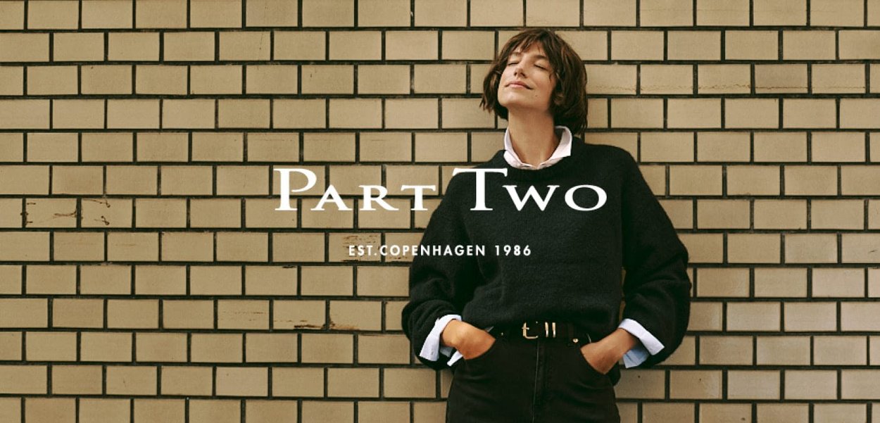 Schwarzer gestrickter Pullover mit weiten Ärmeln, kombiniert mit hoch taillierten dunklen Hosen und einem weißen Hemdkragen. Textüberlagerung: "Part Two EST. COPENHAGEN 1986."