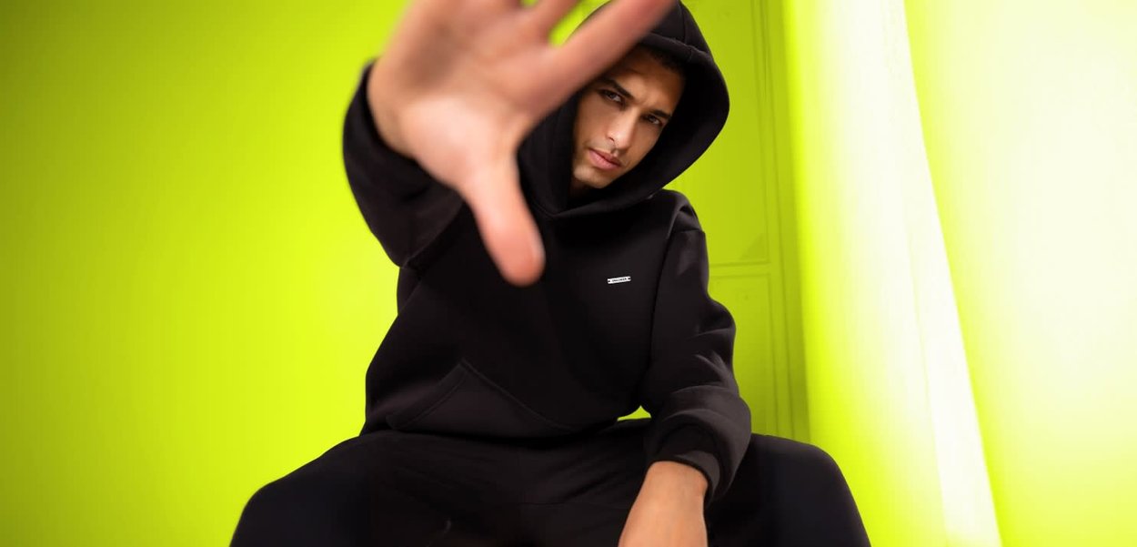 Schwarzer Kapuzenpullover mit einer Fronttasche, versehen mit einem kleinen weißen Logo. Glatte Textur, getragen mit passenden schwarzen Hosen. Grüner Hintergrund.