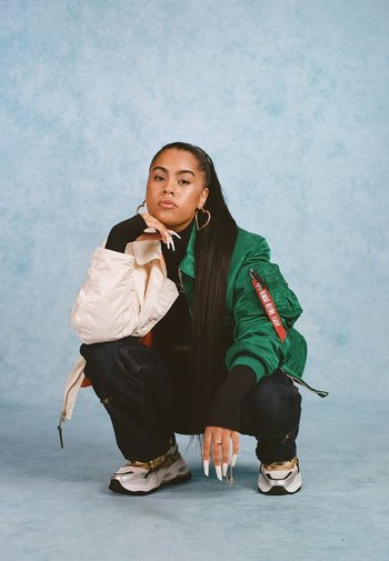 Groene en crèmekleurige bomberjack, zwarte trui, donkere jeans, gouden sneakers met chunky ontwerp, lange nagels en creolen.