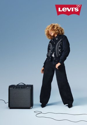 Veste noire courte avec broderie blanche, associée à un pantalon noir à taille haute et jambes larges. Amplificateur noir avec des boutons et un câble au sol.