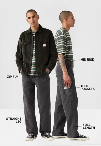 Chaqueta de pana negra, camiseta de rayas verdes y blancas, y pantalones grises de pierna recta con bolsillos para herramientas, cremallera y de longitud completa.