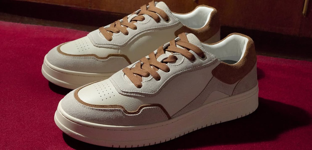 Weiss-beige Sneaker mit strukturiertem Obermaterial aus Wildleder und Leder, braunen Schuhbändern, perforierter Spitze und mehrlagigem Design, liegend auf rotem Hintergrund.