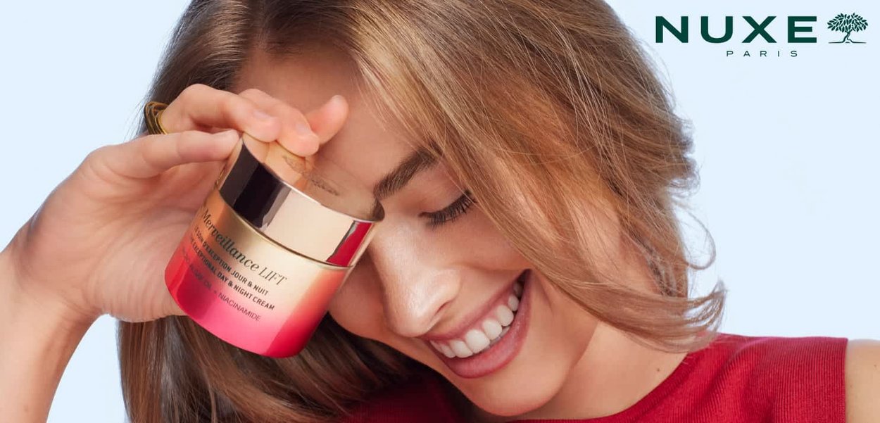 Donna che sorride mentre tiene la crema viso giorno e notte NUXE Paris Revelatrice LIFT accostata alla guancia.