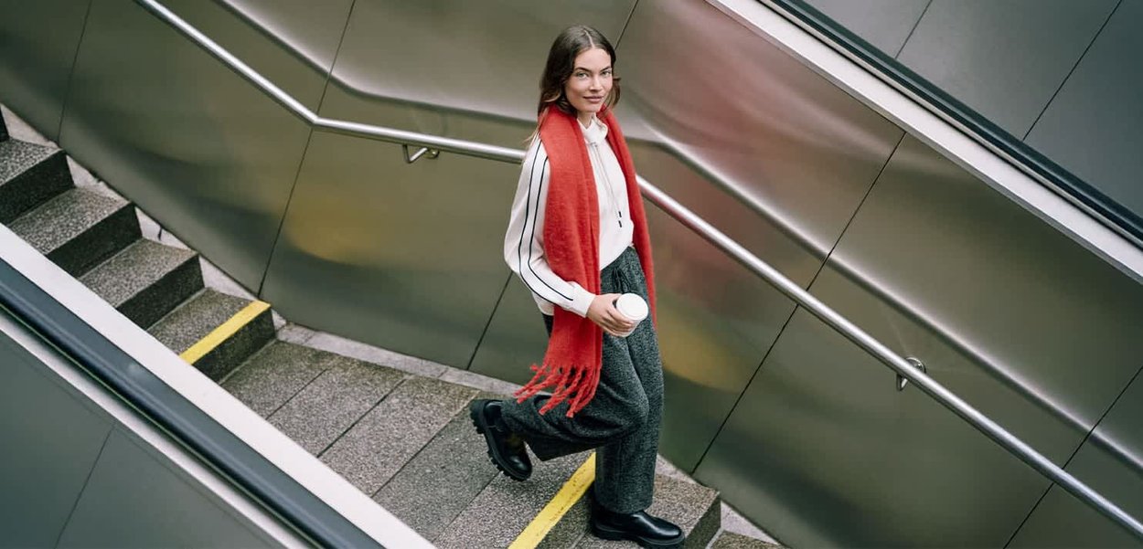 Écharpe rouge, chemise blanche à rayures noires, pantalon gris, tenant une tasse de café, descendant des escaliers avec une rampe en acier inoxydable.