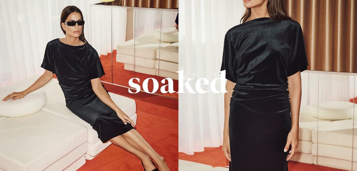 Schwarzes, satinartiges Kleid mit entspannter Silhouette, umgeschlagenen Ärmeln und gerafftem Stoff an der Taille. Model sitzt im Hintergrund vor minimalistischer Möbeln.