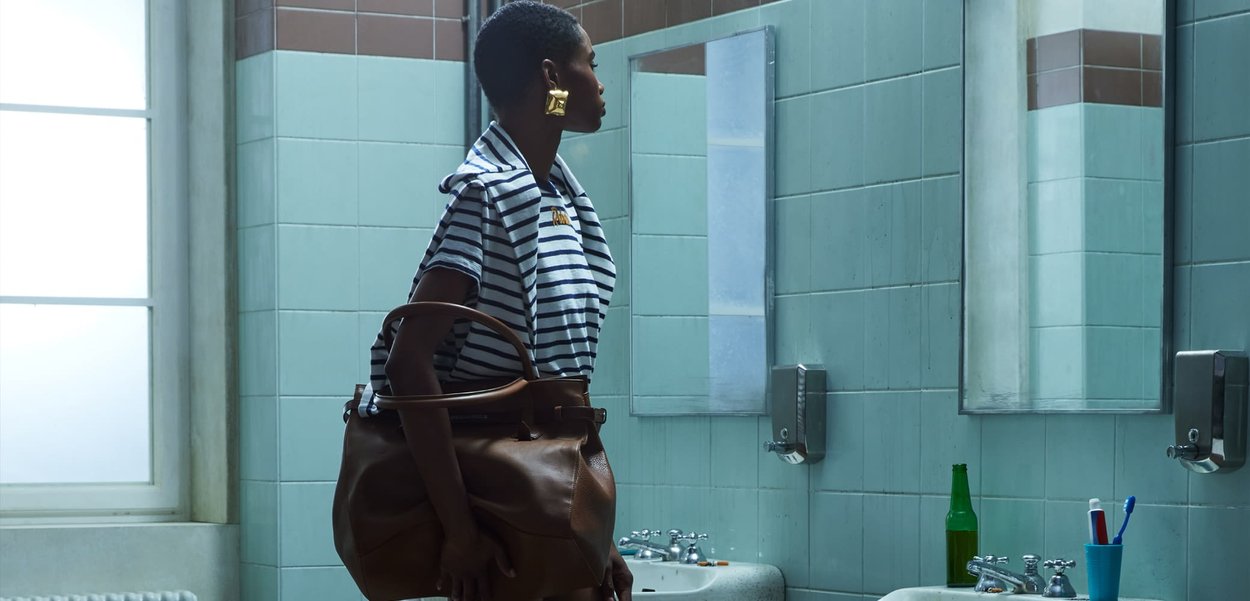 Mujer con cabello corto, camiseta a rayas, grandes pendientes y bolso marrón, de pie en un baño público con azulejos, frente a un espejo sobre dos lavabos.