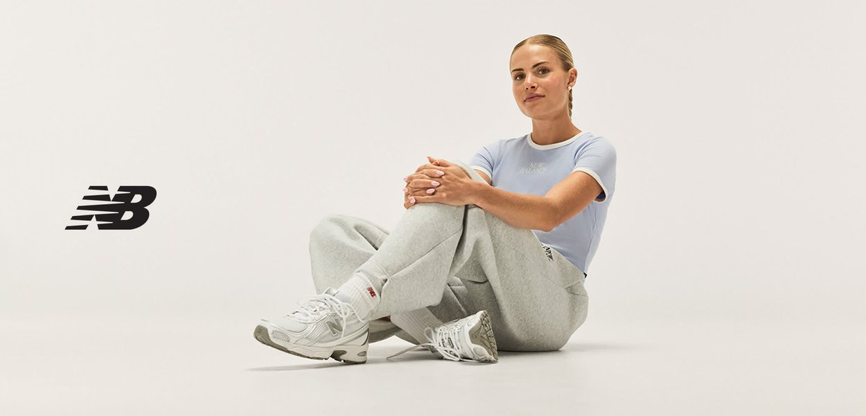 Femme assise par terre portant une chemise bleu clair, un pantalon de survêtement gris et des baskets blanches New Balance avec le logo sur le côté gauche.