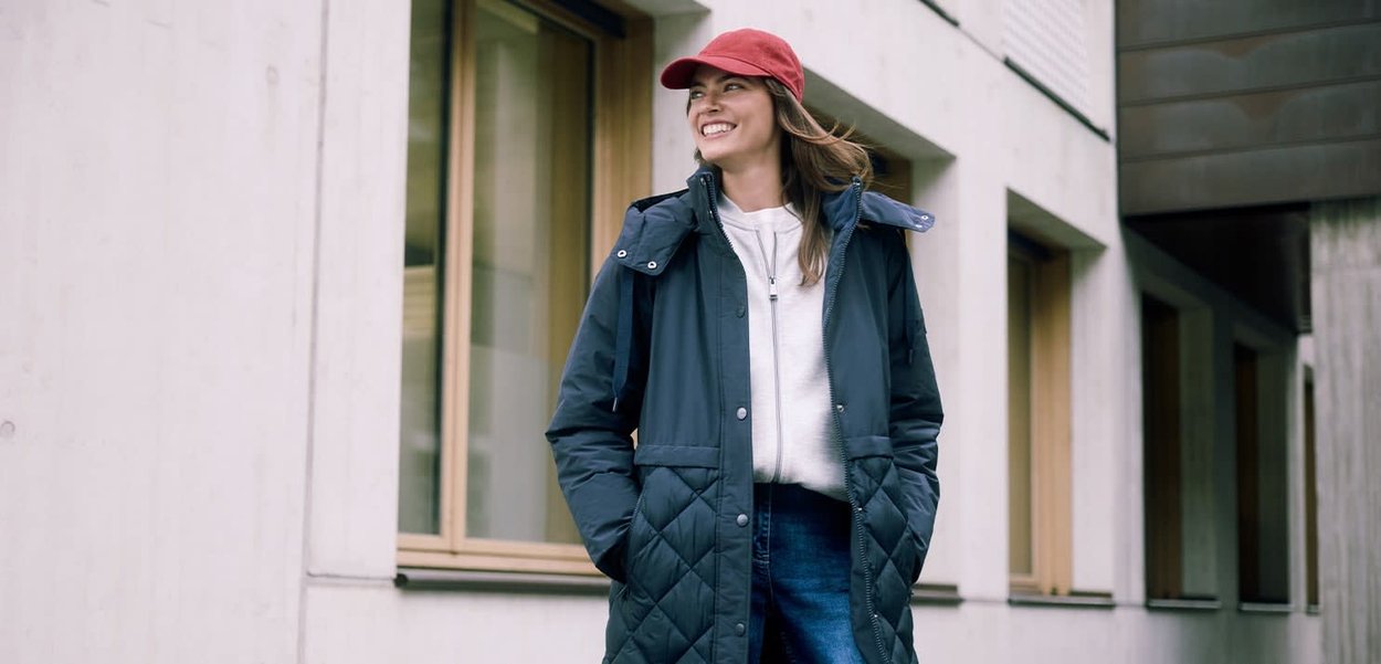 Ung kvinde iført en rød kasket, mørk quiltet jakke, hvid trøje med lynlås og blå jeans, stående nær et moderne bygningsvindue.