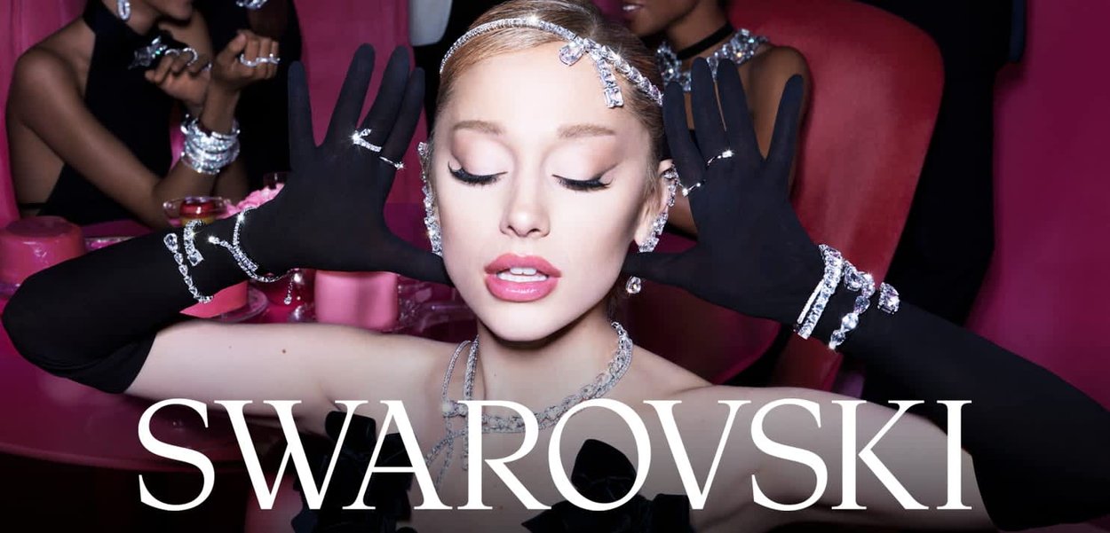 Swarovski sieraden tonen sprankelende kristallen op een model, die zwarte handschoenen en een ketting draagt. De achtergrond is roze met zachte meubels.