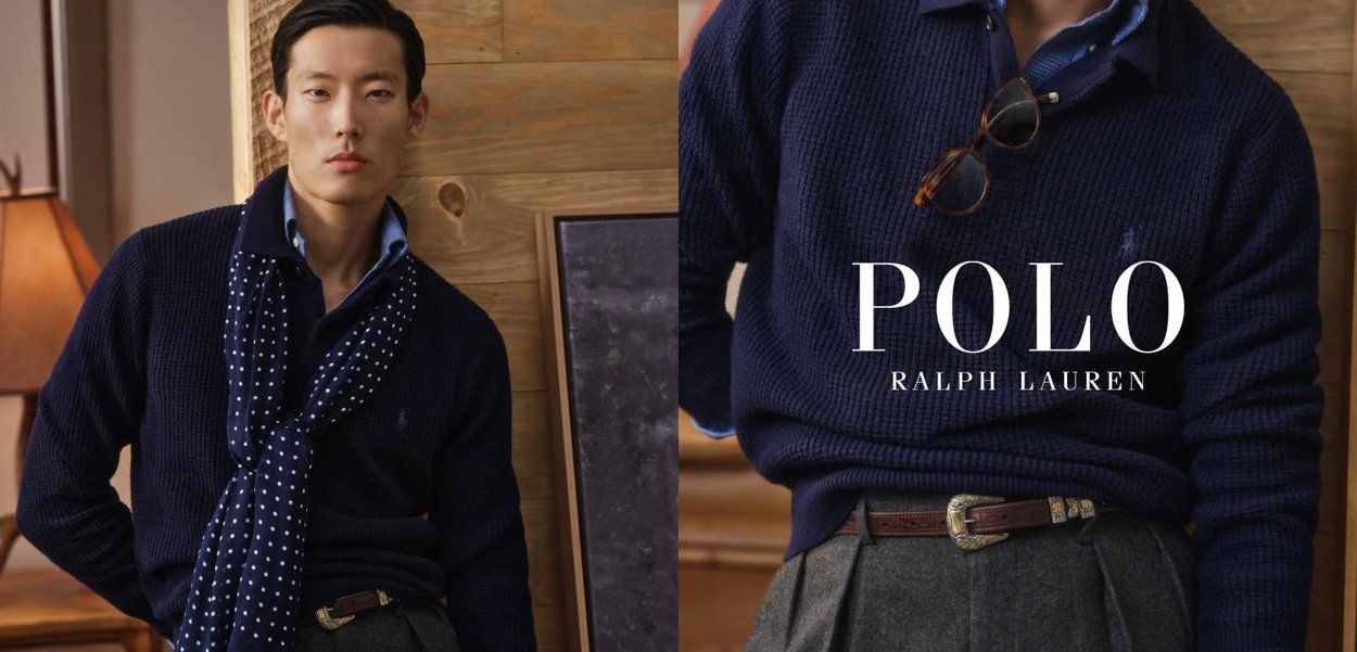 Uomo con maglione blu navy a maglia, sciarpa a pois, pantaloni grigi e cintura marrone, con occhiali appesi al colletto del maglione.