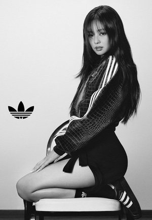 Kobieta klęcząca na krześle, ubrana w teksturowaną kurtkę Adidas, czarną spódnicę, skarpetki i trampki z paskami Adidas w monochromatycznym otoczeniu.