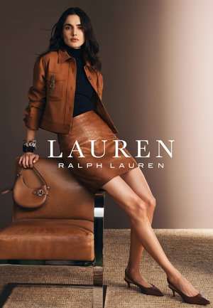 Giacca di pelle marrone su un top nero, abbinata a una gonna a tubino marrone texturizzata. Modella seduta su una sedia metallica elegante, con una borsa in mano.